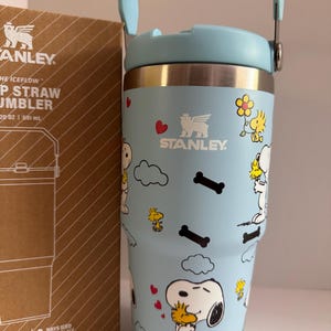 May include: A light blue Stanley Iceflow Flip Straw Tumbler with a cartoon Snoopy and Woodstock design. The tumbler has a handle and a lid. The box next to it has the Stanley logo and product information. The tumbler holds 20 oz (591 ml).