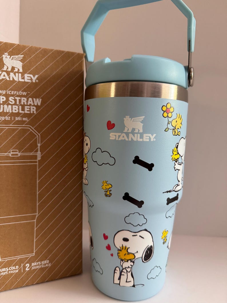 May include: A light blue Stanley Iceflow Flip Straw Tumbler with a cartoon Snoopy and Woodstock design. The tumbler has a handle and a lid. The box next to it has the Stanley logo and product information. The tumbler holds 20 oz (591 ml).