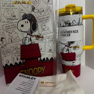 May include: A white Stanley Quencher H2.0 tumbler with a yellow handle and lid, featuring a Snoopy graphic. It includes a clear straw, a white branded dust bag, and a care instruction card, all presented in a matching Snoopy-themed box.