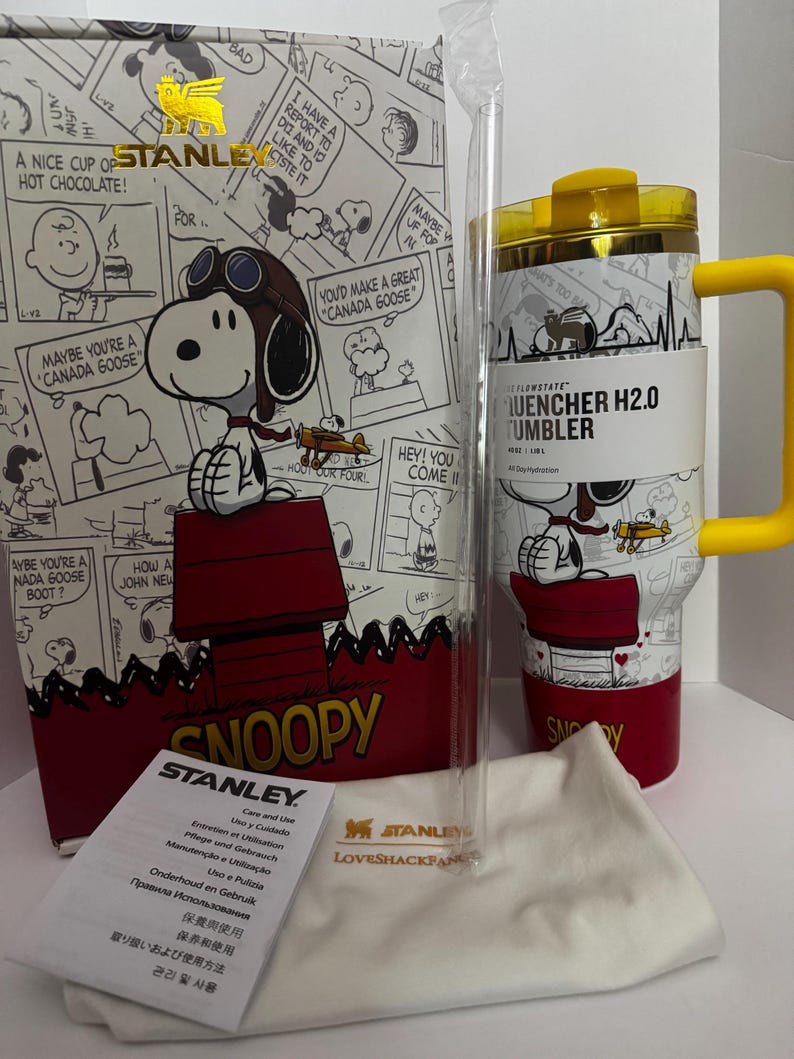 May include: A white Stanley Quencher H2.0 tumbler with a yellow handle and lid, featuring a Snoopy graphic. It includes a clear straw, a white branded dust bag, and a care instruction card, all presented in a matching Snoopy-themed box.