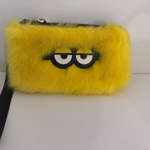 May include: A yellow, rectangular, furry clutch with a black wrist strap. The clutch features a pair of cartoon eyes with white irises and black outlines. The zipper is gold-colored.