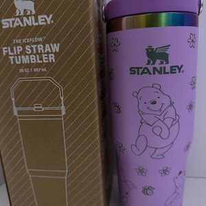 Puede incluir: Vaso Stanley Iceflow Flip Straw morado, 887 ml, con dise&ntilde;o de Winnie the Pooh. Tiene pajita abatible y asa. La caja contigua muestra el logotipo de Stanley y detalles del producto.