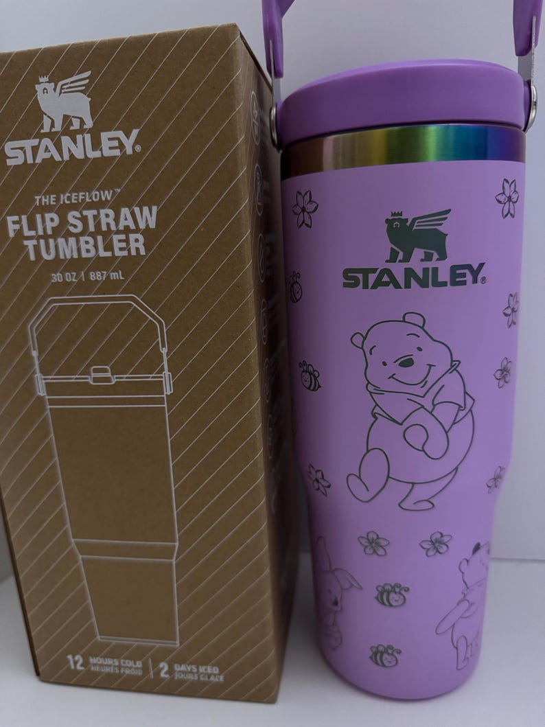 Puede incluir: Vaso Stanley Iceflow Flip Straw morado, 887 ml, con dise&ntilde;o de Winnie the Pooh. Tiene pajita abatible y asa. La caja contigua muestra el logotipo de Stanley y detalles del producto.