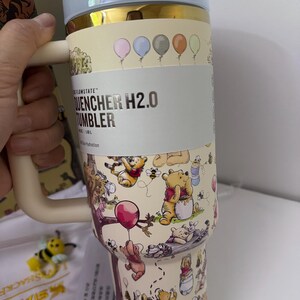 K&ouml;nnte beinhalten: Ein beigefarbener Isolierbecher mit Winnie-the-Pooh-Design. Der Becher hat einen Griff, einen hellblauen Deckel und goldfarbene Akzente. Der Text "FLOWSTATE QUENCHER H2.0 TUMBLER" ist auf der Seite aufgedruckt. Der Becher ist mit Zeichentrickfiguren und Luftballons verziert.