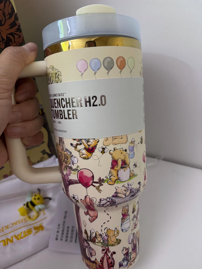 K&ouml;nnte beinhalten: Ein beigefarbener Isolierbecher mit Winnie-the-Pooh-Design. Der Becher hat einen Griff, einen hellblauen Deckel und goldfarbene Akzente. Der Text "FLOWSTATE QUENCHER H2.0 TUMBLER" ist auf der Seite aufgedruckt. Der Becher ist mit Zeichentrickfiguren und Luftballons verziert.