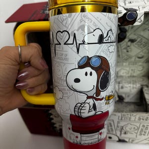 May include: A white and red stainless steel tumbler featuring a graphic of Snoopy as a flying ace with goggles, a heart-shaped heartbeat line, and comic strip panels. The tumbler has a yellow handle, a yellow lid, and the word SNOOPY in yellow letters.