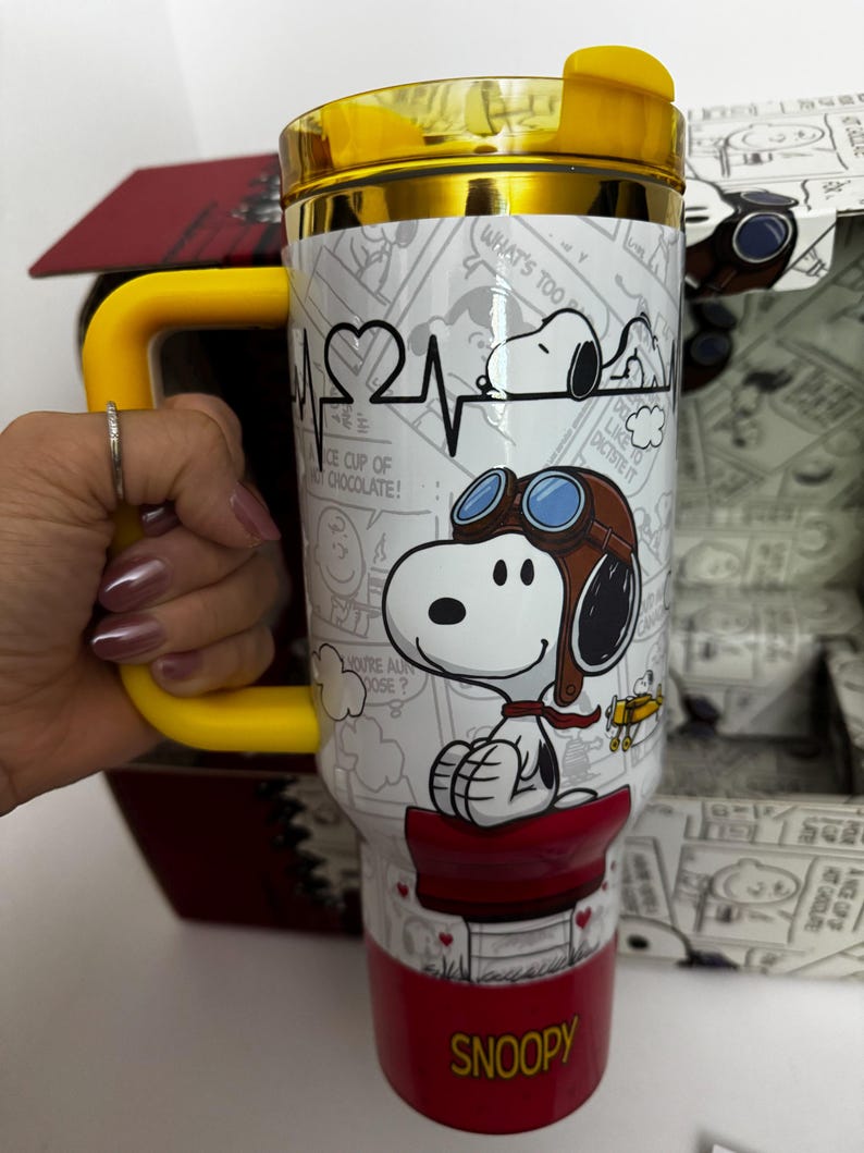 May include: A white and red stainless steel tumbler featuring a graphic of Snoopy as a flying ace with goggles, a heart-shaped heartbeat line, and comic strip panels. The tumbler has a yellow handle, a yellow lid, and the word SNOOPY in yellow letters.