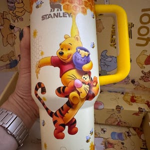 May include: A Stanley insulated tumbler featuring Winnie the Pooh and Tigger. The tumbler has a yellow handle and lid, with a cream-colored body decorated with honeycombs, bees, and the characters. The Stanley logo is visible.