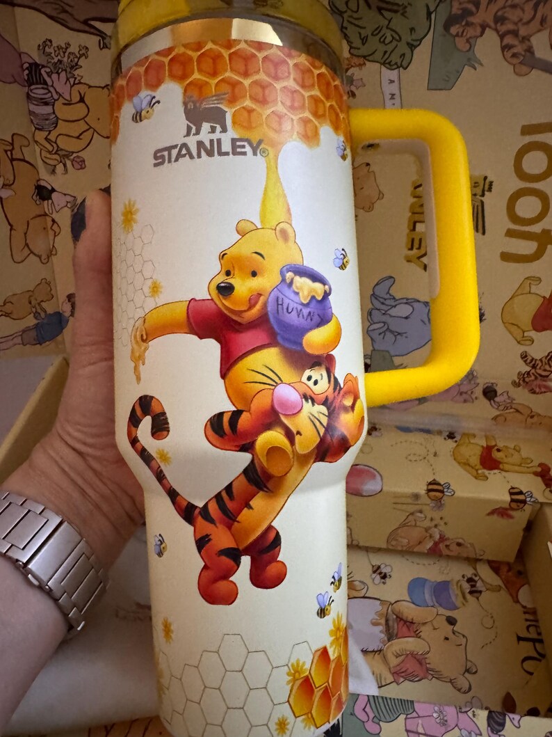 May include: A Stanley insulated tumbler featuring Winnie the Pooh and Tigger. The tumbler has a yellow handle and lid, with a cream-colored body decorated with honeycombs, bees, and the characters. The Stanley logo is visible.