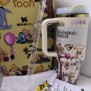 K&ouml;nnte beinhalten: Ein Stanley Quencher H2.0 Becher im Winnie-the-Pooh-Design mit Strohhalm, einer Box und einem Bienen-Charm. Der Becher zeigt Illustrationen von Winnie-the-Pooh-Figuren und Ballons. Auf der Box steht "Winnie the Pooh" und "Stanley".