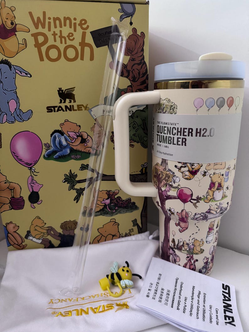 K&ouml;nnte beinhalten: Ein Stanley Quencher H2.0 Becher im Winnie-the-Pooh-Design mit Strohhalm, einer Box und einem Bienen-Charm. Der Becher zeigt Illustrationen von Winnie-the-Pooh-Figuren und Ballons. Auf der Box steht "Winnie the Pooh" und "Stanley".