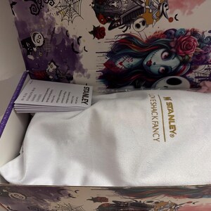 Puede incluir: Caja abierta con una bolsa blanca con cord&oacute;n en el interior, con el texto dorado "STANLEY LOVESHACK FANCY". El interior de la caja tiene una decoraci&oacute;n g&oacute;tica, con ilustraciones de personajes y elementos florales.
