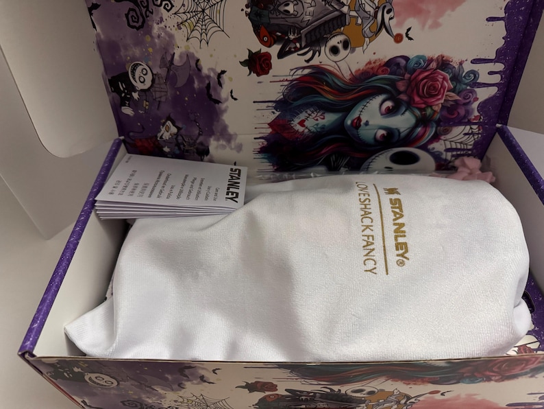 Puede incluir: Caja abierta con una bolsa blanca con cord&oacute;n en el interior, con el texto dorado "STANLEY LOVESHACK FANCY". El interior de la caja tiene una decoraci&oacute;n g&oacute;tica, con ilustraciones de personajes y elementos florales.