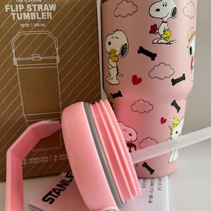 May include: A pink Stanley The Iceflow Flip Straw Tumbler, 20 oz, featuring Snoopy and Woodstock graphics. The tumbler includes a pink handle and lid, and a clear straw. The box is visible in the background.