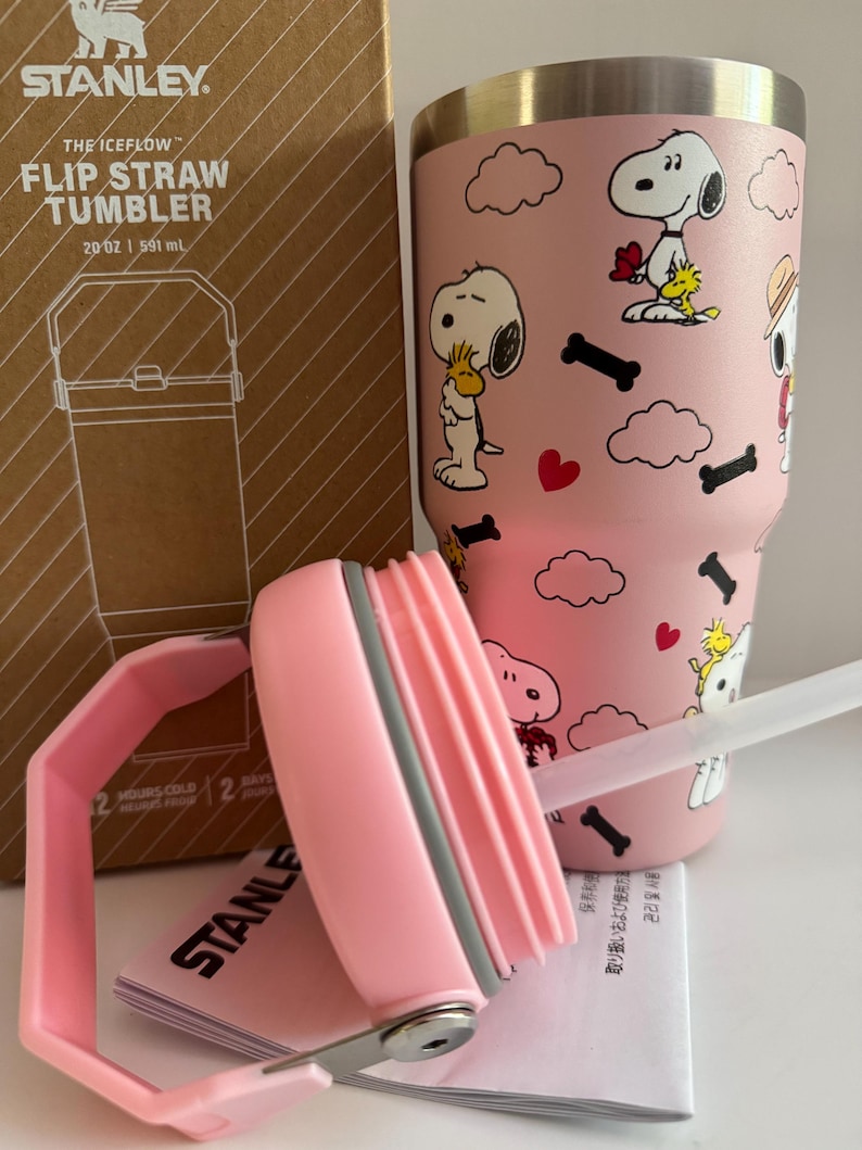 May include: A pink Stanley The Iceflow Flip Straw Tumbler, 20 oz, featuring Snoopy and Woodstock graphics. The tumbler includes a pink handle and lid, and a clear straw. The box is visible in the background.