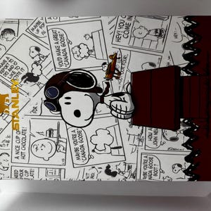 May include: A rectangular box featuring a black and white Peanuts comic strip pattern with Snoopy wearing aviator goggles. The box includes a dark red section with the word SNOOPY in gold lettering and a gold Stanley logo on the left side.