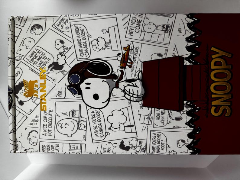 May include: A rectangular box featuring a black and white Peanuts comic strip pattern with Snoopy wearing aviator goggles. The box includes a dark red section with the word SNOOPY in gold lettering and a gold Stanley logo on the left side.