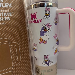 Op de afbeelding: Witte Stanley Quencher H2.0 FlowState-beker met een roze handvat en Daisy Duck cartoonafbeeldingen. De beker heeft een zilveren rand en een doorzichtig deksel. De doos ernaast heeft het Stanley-logo en de tekst "Quencher H2.0 FlowState Tumbler".