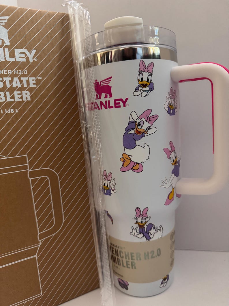 Op de afbeelding: Witte Stanley Quencher H2.0 FlowState-beker met een roze handvat en Daisy Duck cartoonafbeeldingen. De beker heeft een zilveren rand en een doorzichtig deksel. De doos ernaast heeft het Stanley-logo en de tekst "Quencher H2.0 FlowState Tumbler".