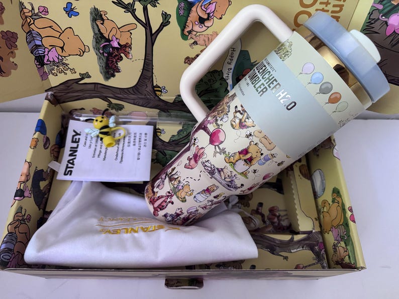 K&ouml;nnte beinhalten: Ein Stanley Quencher H2.0 Becher mit Winnie Puuh-Design, hellblauem Deckel und Griff. Der Becher befindet sich in einer dekorativen Box mit Winnie Puuh-Illustrationen, einer kleinen Bienenfigur und einem wei&szlig;en Kordelzugbeutel.