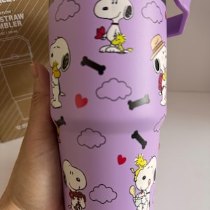 May include: A purple Stanley FlowStraw tumbler featuring Snoopy and Woodstock cartoon graphics. The tumbler has a purple handle and lid, with a silver hinge. The design includes clouds, hearts, and bone shapes.