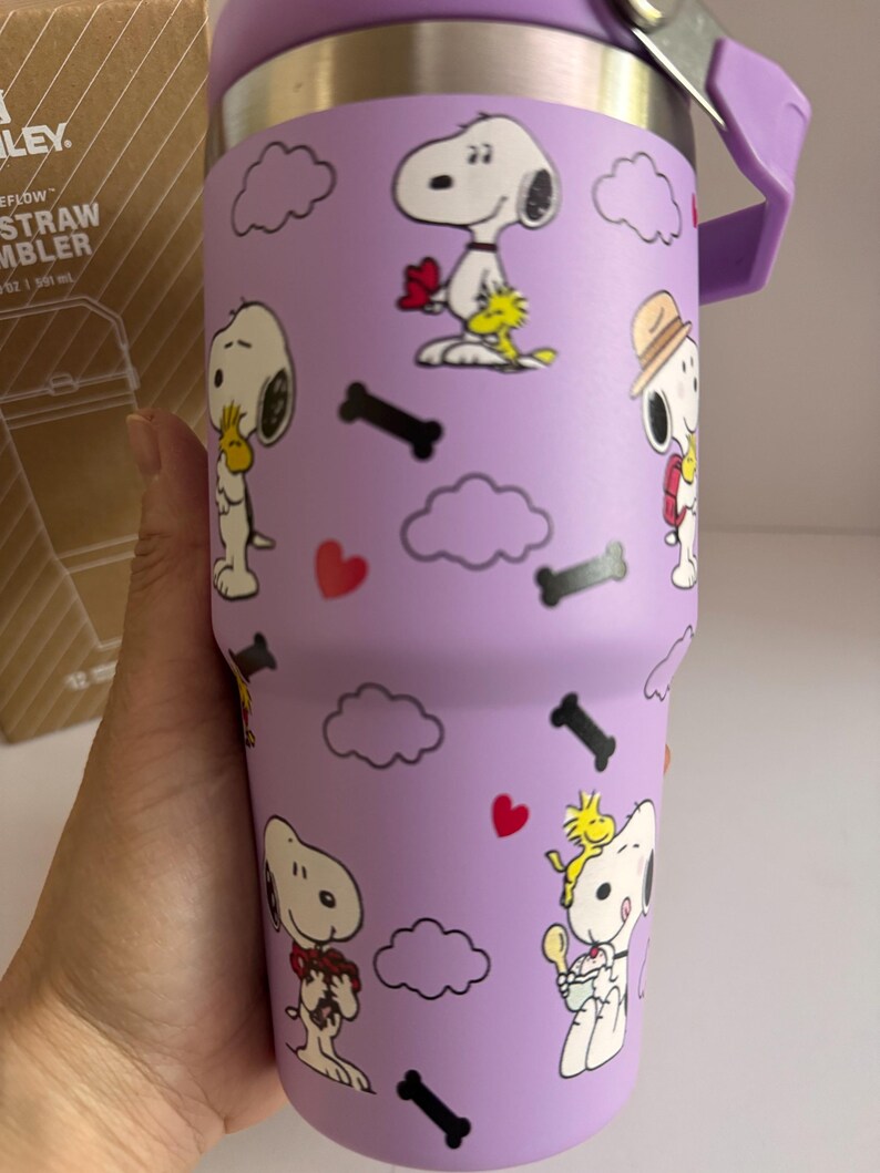 May include: A purple Stanley FlowStraw tumbler featuring Snoopy and Woodstock cartoon graphics. The tumbler has a purple handle and lid, with a silver hinge. The design includes clouds, hearts, and bone shapes.
