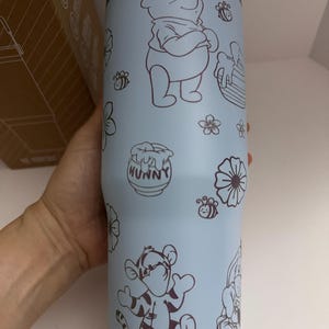 Puede incluir: Vaso t&eacute;rmico azul claro con ilustraciones de Winnie the Pooh, Tigger, abejas, flores y tarros de miel. El vaso tiene un asa azul claro y un borde plateado. La palabra "HUNNY" est&aacute; en el tarro de miel.