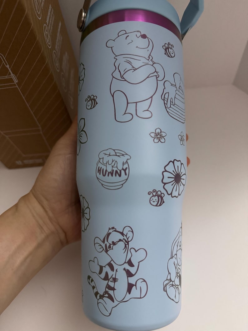 Puede incluir: Vaso t&eacute;rmico azul claro con ilustraciones de Winnie the Pooh, Tigger, abejas, flores y tarros de miel. El vaso tiene un asa azul claro y un borde plateado. La palabra "HUNNY" est&aacute; en el tarro de miel.