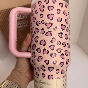 May include: A pink Stanley Quencher tumbler with a pink handle and lid. The tumbler features a leopard print pattern of pink hearts on a light pink background. The tumbler has a double-wall vacuum insulation and is car cup holder compatible.