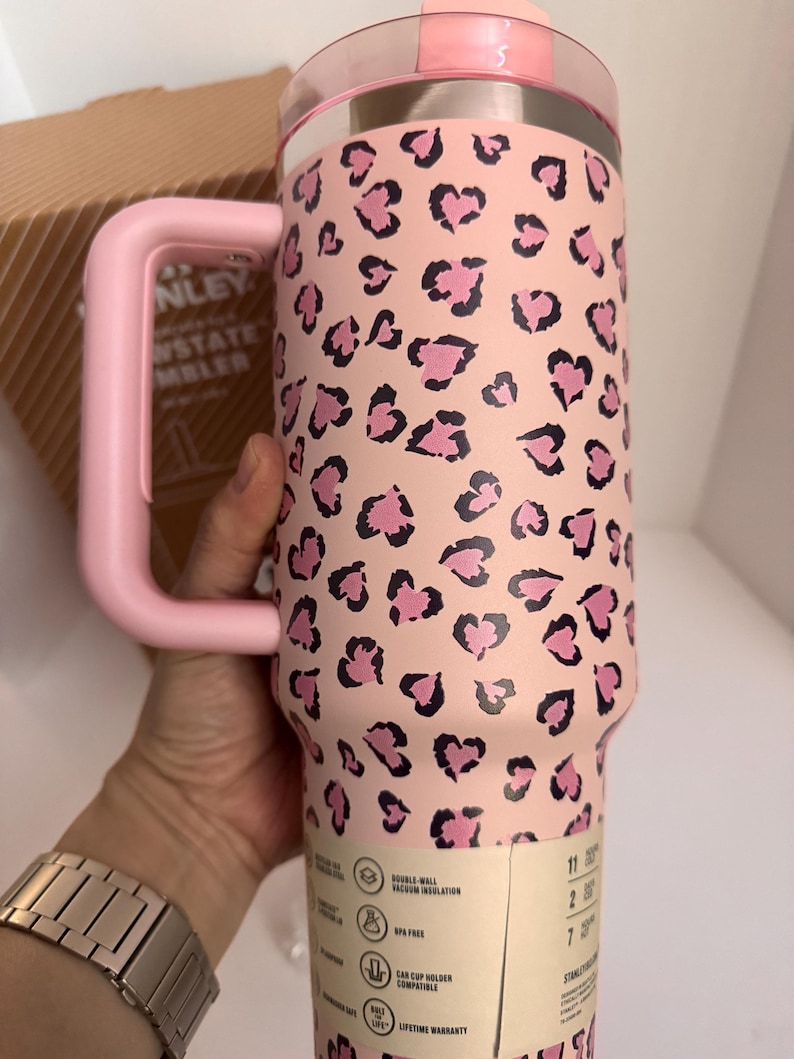 May include: A pink Stanley Quencher tumbler with a pink handle and lid. The tumbler features a leopard print pattern of pink hearts on a light pink background. The tumbler has a double-wall vacuum insulation and is car cup holder compatible.