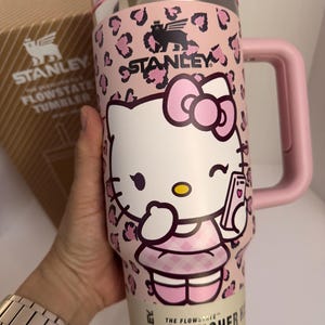 May include: A pink Stanley Flowstate Quencher Tumbler featuring a Hello Kitty design. The tumbler has a pink leopard print pattern and a pink handle. The text "THE FLOWSTATE QUENCHER TUMBLER" is printed on the bottom. It holds 40 oz.