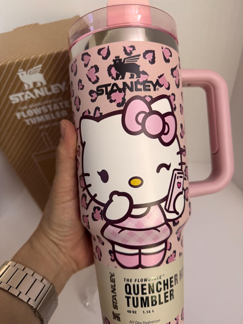 May include: A pink Stanley Flowstate Quencher Tumbler featuring a Hello Kitty design. The tumbler has a pink leopard print pattern and a pink handle. The text "THE FLOWSTATE QUENCHER TUMBLER" is printed on the bottom. It holds 40 oz.