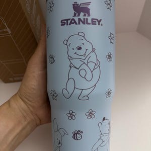 Puede incluir: Vaso Stanley azul claro con ilustraciones de Winnie the Pooh y sus amigos. El vaso tiene un borde y accesorios de asa morados. El dise&ntilde;o incluye a Winnie the Pooh, un conejo, abejas y flores. El logotipo de Stanley tambi&eacute;n es visible.