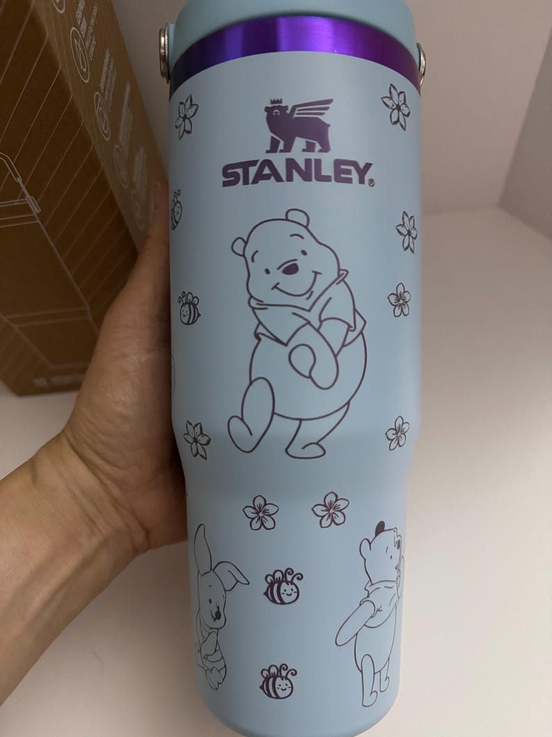 Puede incluir: Vaso Stanley azul claro con ilustraciones de Winnie the Pooh y sus amigos. El vaso tiene un borde y accesorios de asa morados. El dise&ntilde;o incluye a Winnie the Pooh, un conejo, abejas y flores. El logotipo de Stanley tambi&eacute;n es visible.