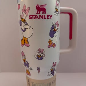 Op de afbeelding: Witte Stanley Quencher H2.0 Flowstate tumbler met Daisy Duck cartoonafbeeldingen. De tumbler heeft een roze handvat en accenten, een zilveren rand en het Stanley-logo. Ontworpen voor de hele dag hydratatie. Inhoud: 1,18 L.