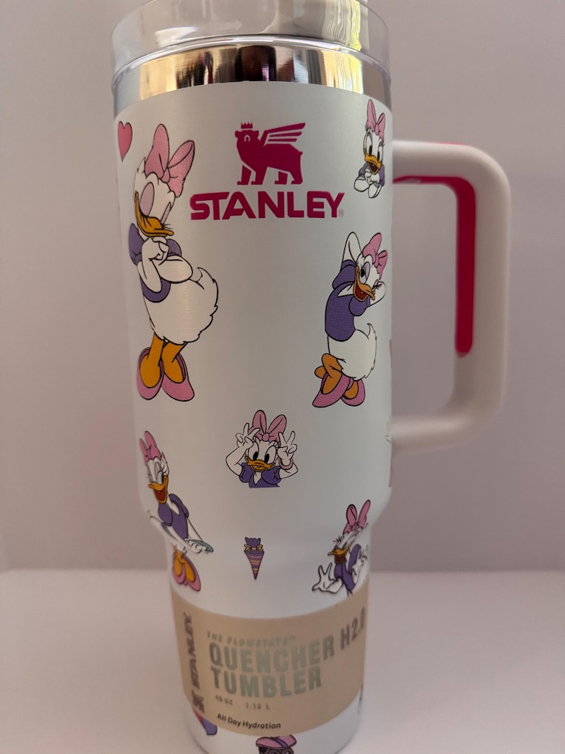 Op de afbeelding: Witte Stanley Quencher H2.0 Flowstate tumbler met Daisy Duck cartoonafbeeldingen. De tumbler heeft een roze handvat en accenten, een zilveren rand en het Stanley-logo. Ontworpen voor de hele dag hydratatie. Inhoud: 1,18 L.