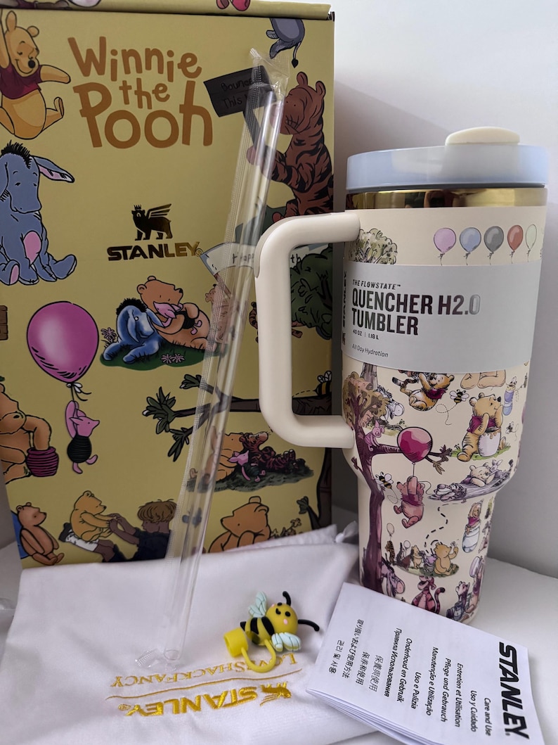 K&ouml;nnte beinhalten: Ein Stanley Quencher H2.0 Becher mit Winnie Puuh-Motiv. Der Becher hat einen hellblauen Deckel und einen wei&szlig;en Griff. Die Box und der Becher zeigen Illustrationen von Winnie Puuh und seinen Freunden. Enth&auml;lt einen Strohhalm und einen bienenf&ouml;rmigen Anh&auml;nger.