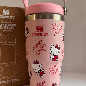 May include: A pink Stanley Iceflow Lip Straw Tumbler featuring a Hello Kitty design. The tumbler has a pink handle and lid, and the body is covered in Hello Kitty illustrations and script. The box next to it has the Stanley logo.
