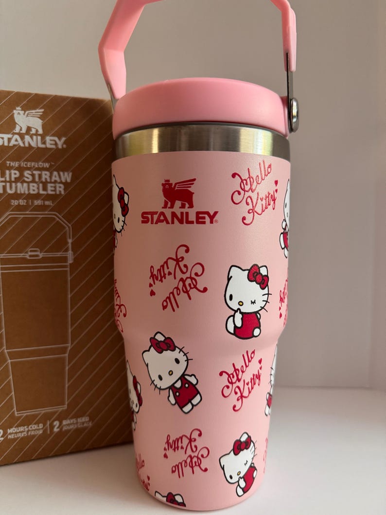 May include: A pink Stanley Iceflow Lip Straw Tumbler featuring a Hello Kitty design. The tumbler has a pink handle and lid, and the body is covered in Hello Kitty illustrations and script. The box next to it has the Stanley logo.