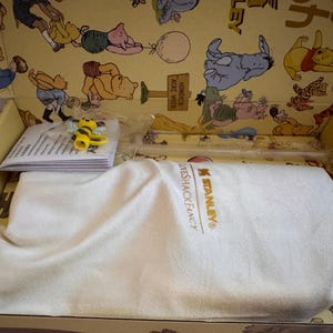 May include: A gift box featuring a Winnie the Pooh theme. Inside, a white cloth covers an item, with a small bee figurine and a stack of papers visible. The box's interior is decorated with Pooh and friends.