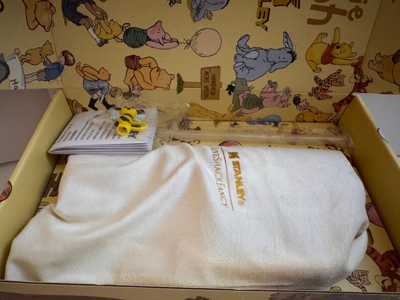 May include: A gift box featuring a Winnie the Pooh theme. Inside, a white cloth covers an item, with a small bee figurine and a stack of papers visible. The box's interior is decorated with Pooh and friends.