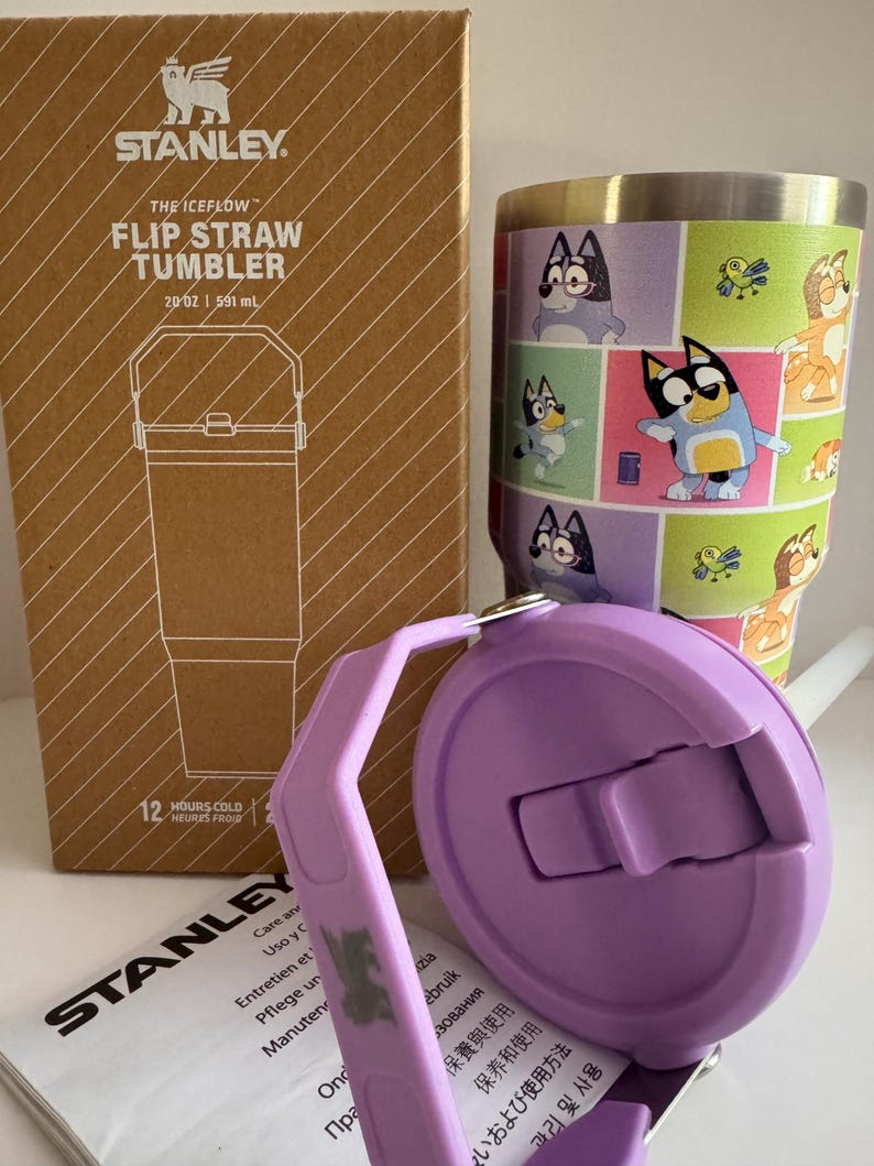 May include: A Stanley The Iceflow Flip Straw Tumbler, 20 oz (591 ml), with a purple lid and a cartoon character design. The tumbler is in its original cardboard box. The lid is open, revealing the straw opening.