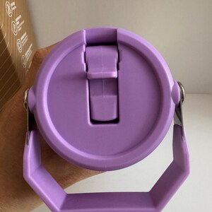May include: A close-up of a lavender-colored water bottle lid with a handle. The lid has a flip-top closure and a silver metal hinge. The handle is a hexagonal shape. The bottle is designed for easy carrying.