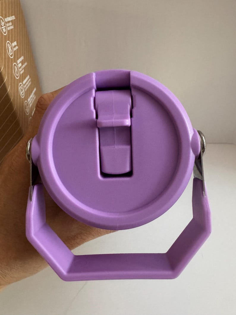 May include: A close-up of a lavender-colored water bottle lid with a handle. The lid has a flip-top closure and a silver metal hinge. The handle is a hexagonal shape. The bottle is designed for easy carrying.