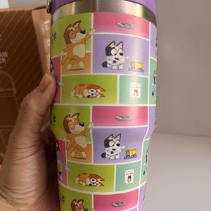May include: A stainless steel tumbler with a purple lid and handle. The tumbler features a colorful cartoon design of dogs in various poses, set against a backdrop of pink, green, and purple squares. The tumbler is held by a hand.