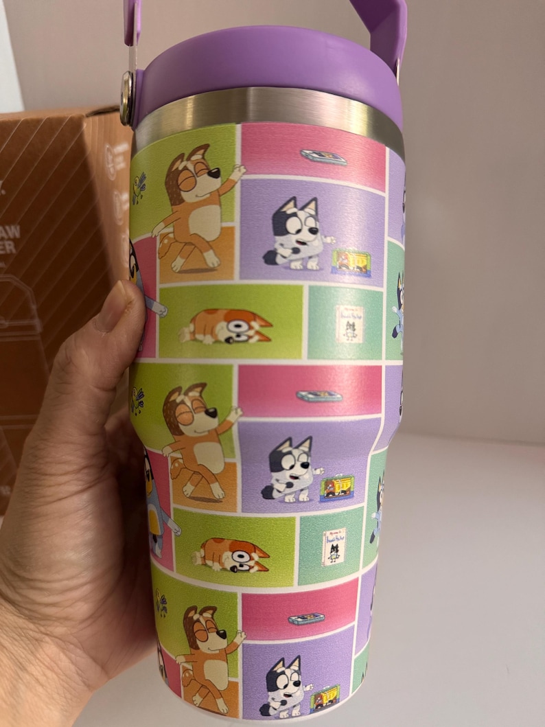 May include: A stainless steel tumbler with a purple lid and handle. The tumbler features a colorful cartoon design of dogs in various poses, set against a backdrop of pink, green, and purple squares. The tumbler is held by a hand.