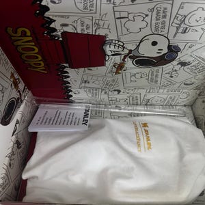 May include: A white fabric drawstring bag with the yellow Stanley logo inside a cardboard box featuring black and white Peanuts comic strip illustrations. The box lid displays the word SNOOPY in yellow and a red doghouse graphic with Snoopy.