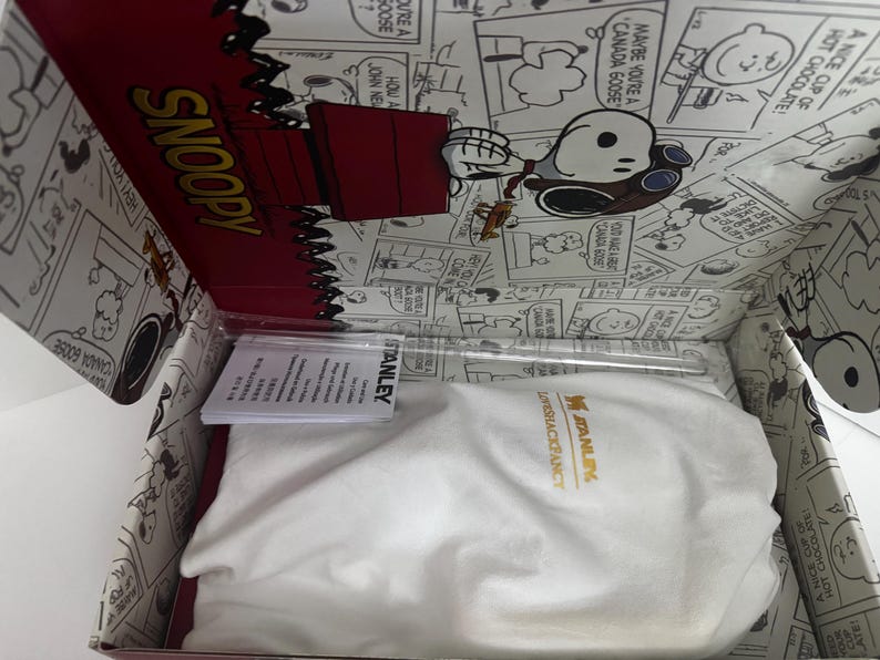 May include: A white fabric drawstring bag with the yellow Stanley logo inside a cardboard box featuring black and white Peanuts comic strip illustrations. The box lid displays the word SNOOPY in yellow and a red doghouse graphic with Snoopy.