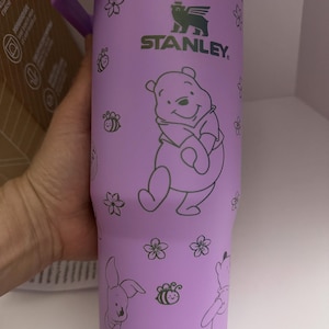 Puede incluir: Vaso Stanley morado con ilustraciones de Winnie the Pooh y sus amigos. El vaso tiene un asa y tapa moradas, con un borde plateado. El dise&ntilde;o incluye Winnie the Pooh, Piglet, abejas y flores. El logotipo de Stanley es visible.