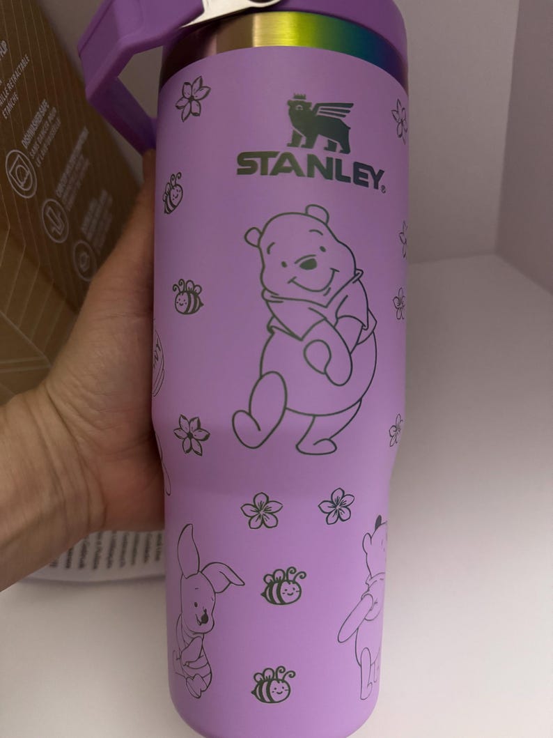 Puede incluir: Vaso Stanley morado con ilustraciones de Winnie the Pooh y sus amigos. El vaso tiene un asa y tapa moradas, con un borde plateado. El dise&ntilde;o incluye Winnie the Pooh, Piglet, abejas y flores. El logotipo de Stanley es visible.